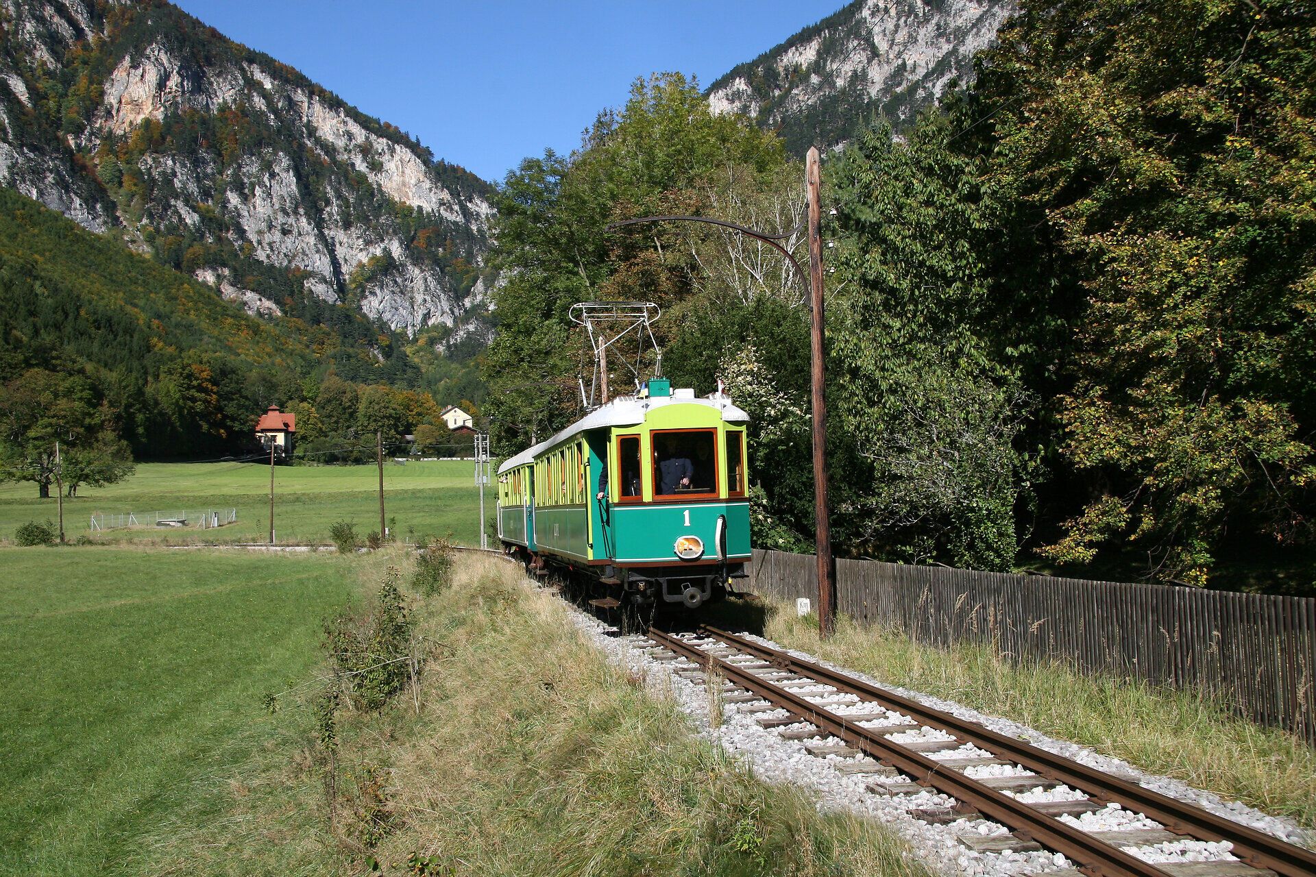 Grün-gelber Schmalspurtriebwagen fährt durch eine Wiesenlandschaft mit Bergen im Hintergrund.