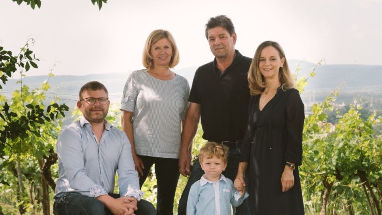 F&uuml;nf Personen posieren in einem Weinberg, darunter ein Kind. Im Hintergrund sind H&uuml;gel und Weinreben zu sehen.