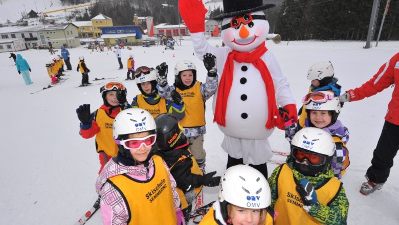 Kinder in Skiausrüstung posieren mit einem Schneemann auf einer Skipiste.