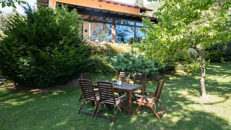 Ein Garten mit Holztisch und Stühlen vor einem Ferienhaus mit großen Fenstern und umgeben von Bäumen und Sträuchern.