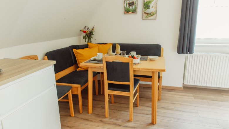 Essbereich mit Holztisch, Stühlen und Eckbank, gedeckt mit Geschirr, Blumenstrauß und Dekoration an der Wand.