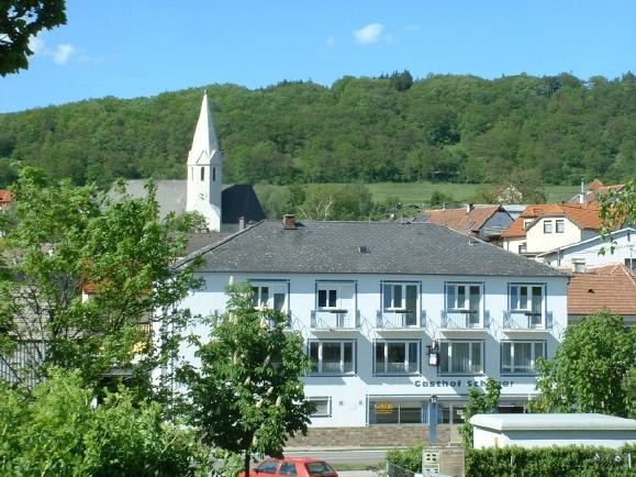 Pension Schauer, © Ingrid Schauer Ein zweistöckiges Gebäude mit der Aufschrift 'Gasthof Schauer' vor einem bewaldeten Hügel und einer Kirche mit spitzem Turm.