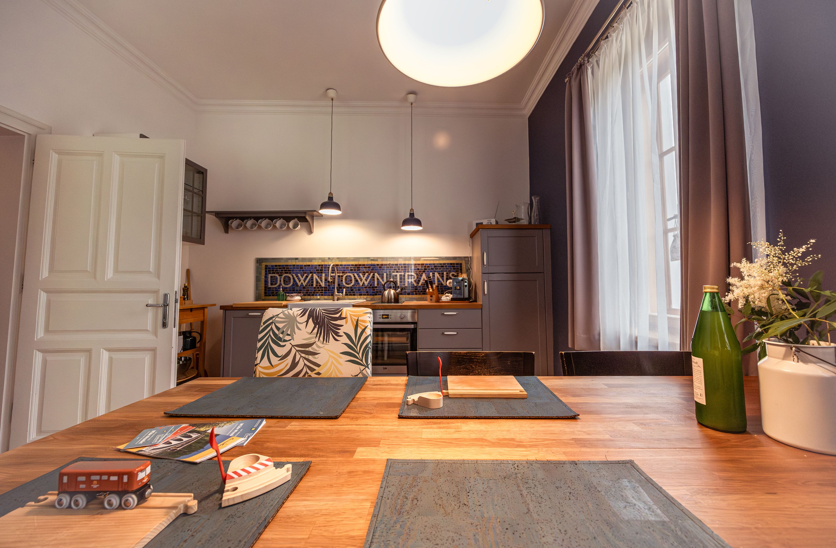 Küche mit Holztisch, Spielzeugzug, Flasche und Blumen auf dem Tisch, moderne Lampen und Küchenschränke im Hintergrund.