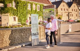 Ein älteres Paar betrachtet eine Informationstafel auf einem Themenweg in einer Stadt mit historischen Gebäuden im Hintergrund.