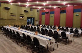 Ein gro&szlig;er Saal mit langen, gedeckten Tischen und schwarzen St&uuml;hlen, bereit f&uuml;r ein Event.