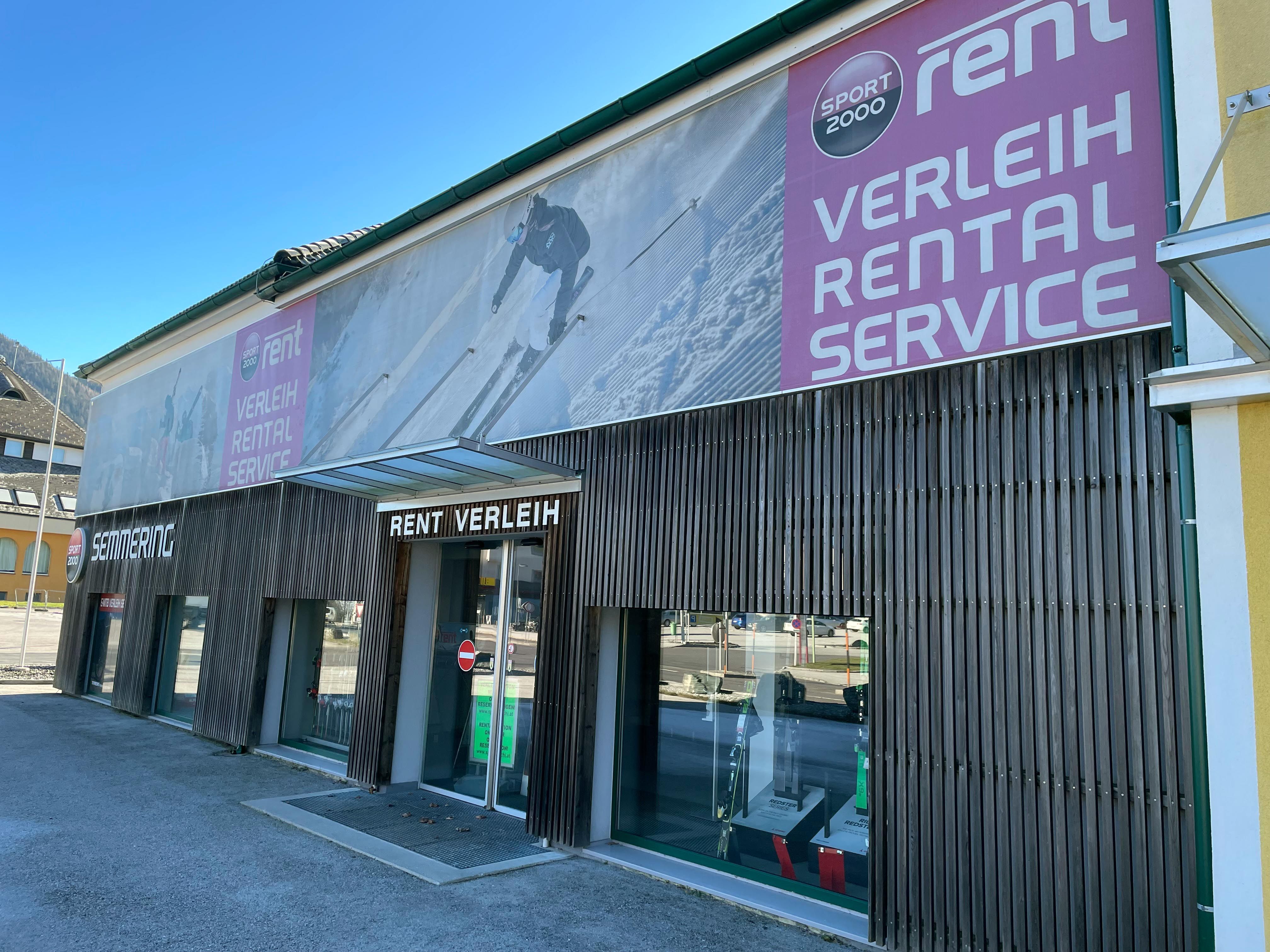 Außenansicht eines Sportverleihgeschäfts mit großen Werbebannern und Schaufenstern.