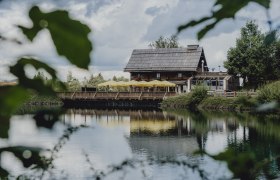Ein rustikales Holzhaus mit gelben Sonnenschirmen spiegelt sich in einem ruhigen See, umgeben von gr&uuml;ner Natur.