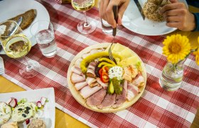 Ein gedeckter Tisch mit einer Platte voller Aufschnitt, K&auml;se und Gem&uuml;se, dazu Wei&szlig;wein und Brot auf einer rot-wei&szlig; karierten Tischdecke.