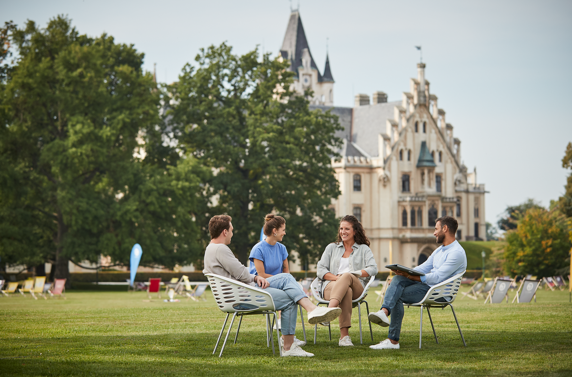 Inmitten einer malerischen Kulisse entspannen sich vier Personen in gemütlichen Stühlen auf einer weitläufigen Wiese. Die sanften Hügel und die beeindruckende Architektur im Hintergrund schaffen eine einladende Atmosphäre, die zum Verweilen und Genießen einlädt.