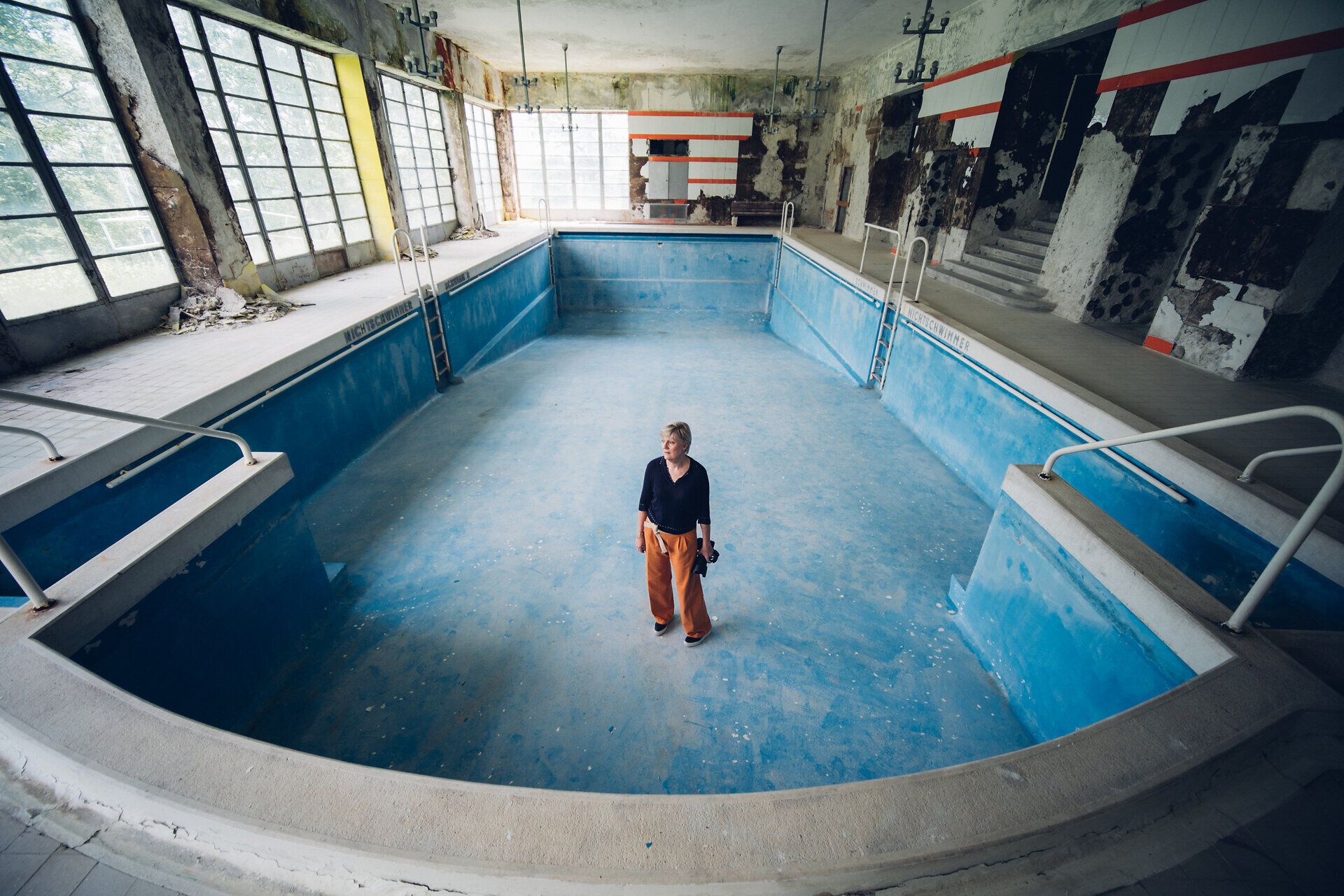 In der stillen, verlassenen Schwimmhalle des Südbahnhotels spürt man die Erinnerungen an vergangene Tage. Das Licht strömt durch die großen Fenster und taucht den Raum in ein sanftes Blau, während die Künstlerin in der Mitte des leeren Beckens steht und die melancholische Schönheit des Ortes einfängt.