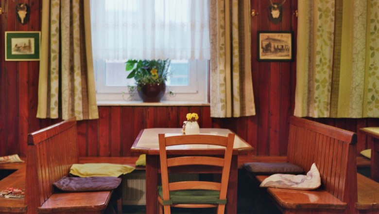 Gemütliche Gaststube mit Holzmöbeln, Tisch, Stühlen und Bank, Fenster mit Vorhängen, Pflanzen und Wanddekorationen.