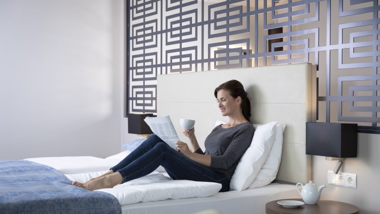Frau entspannt auf einem Bett in einem modernen Hotelzimmer, liest eine Zeitung und h&auml;lt eine Tasse. Dekorative Wand im Hintergrund.