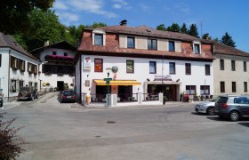 Ein Gebäude mit der Aufschrift 'Dirndlhof' und einer Terrasse mit Sonnenschirm. Autos sind auf dem Parkplatz davor geparkt.