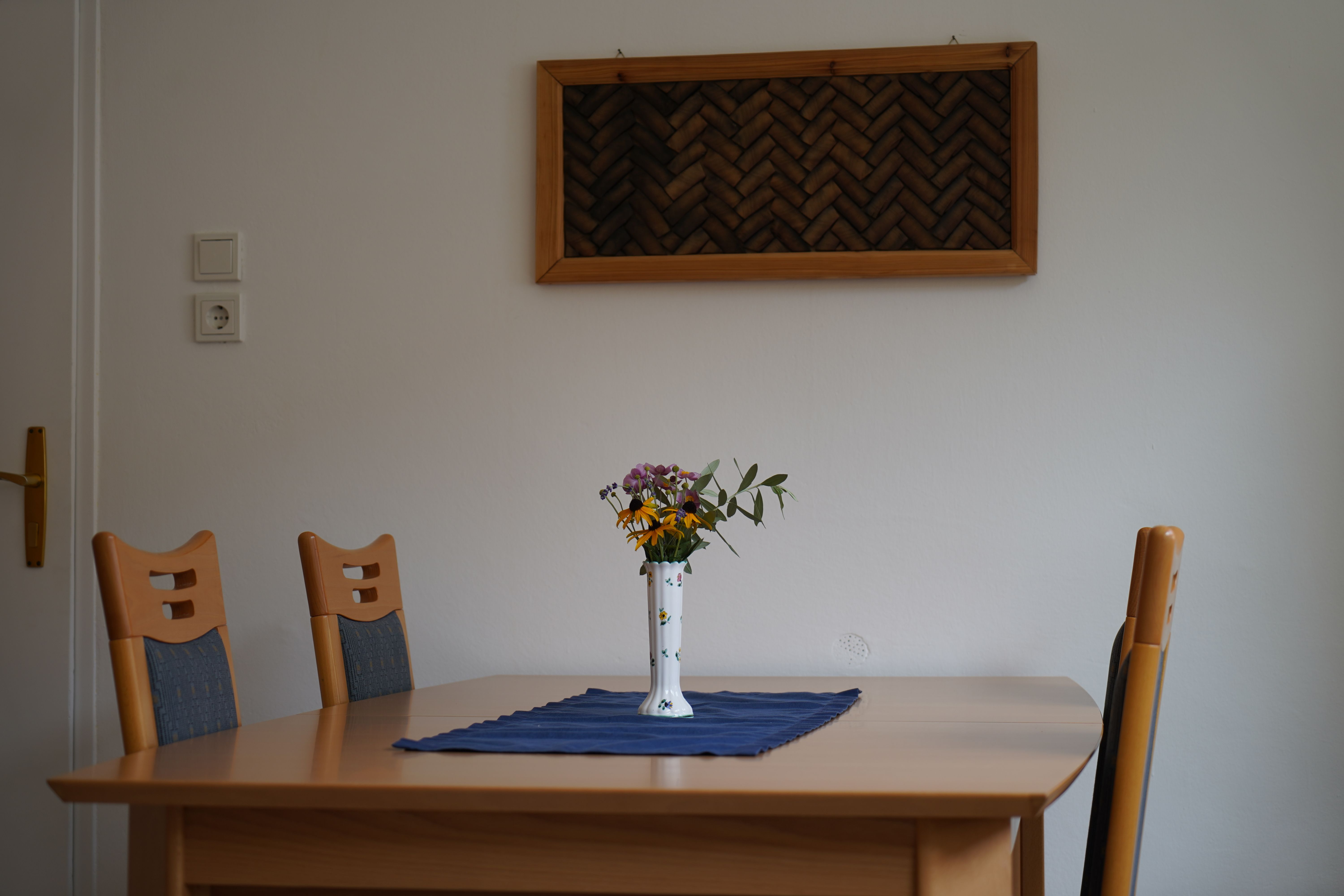 Ein Tisch mit vier Stühlen, einer Vase mit Blumen und einem blauen Tuch in einem Raum mit weißer Wand und einem Wanddekor.