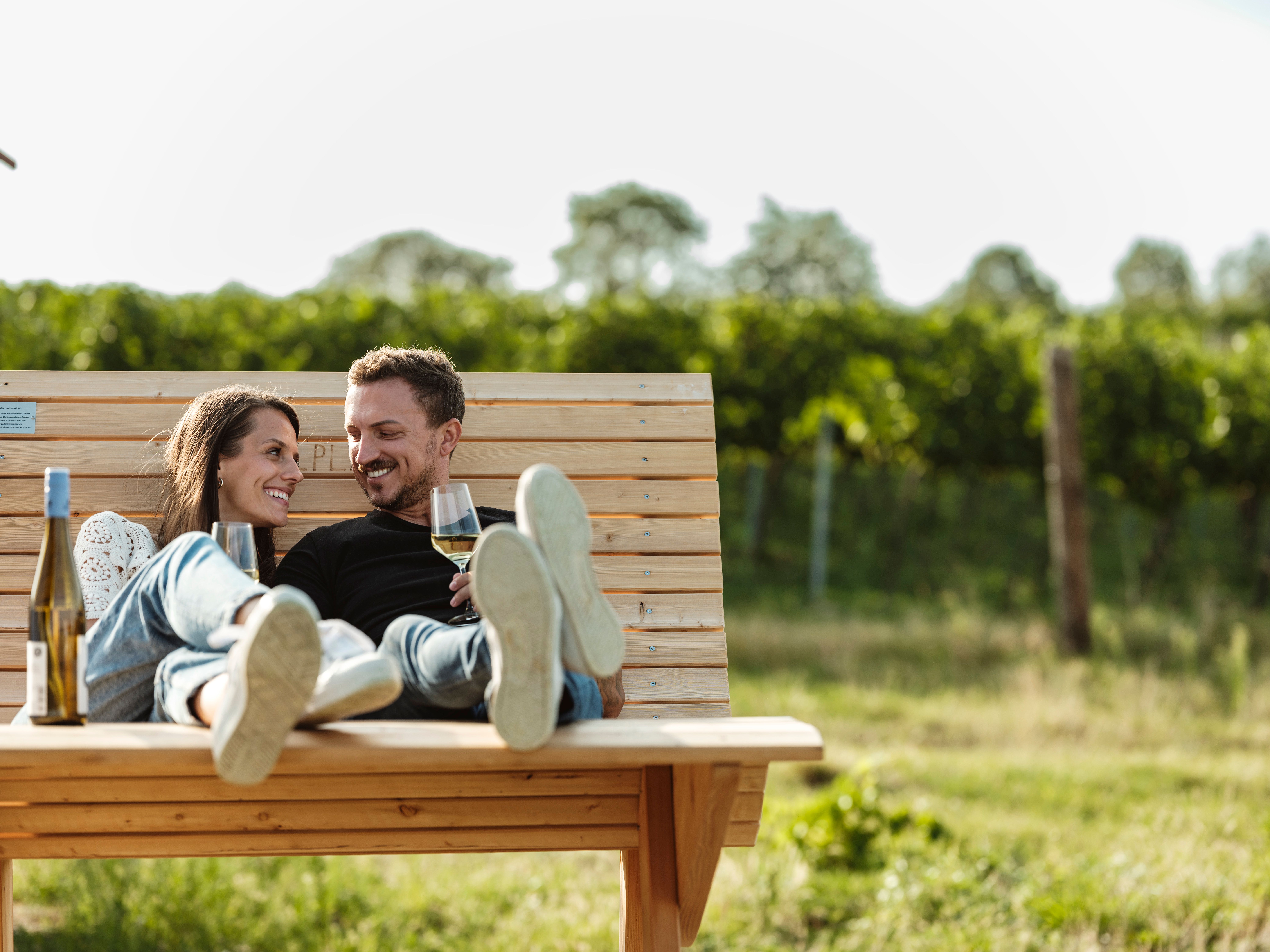 Ein Paar entspannt auf einer Holzbank im Freien mit Weingläsern in der Hand.