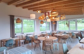 Gem&uuml;tlicher Gastraum mit Holzm&ouml;beln und H&auml;ngelampen, gro&szlig;e Fenster mit Blick ins Gr&uuml;ne.