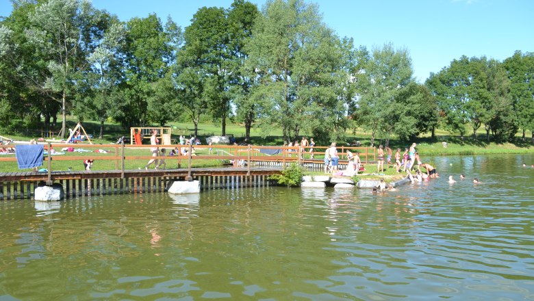 Menschen schwimmen und entspannen an einem Badeteich mit Steg und Spielplatz im Hintergrund.