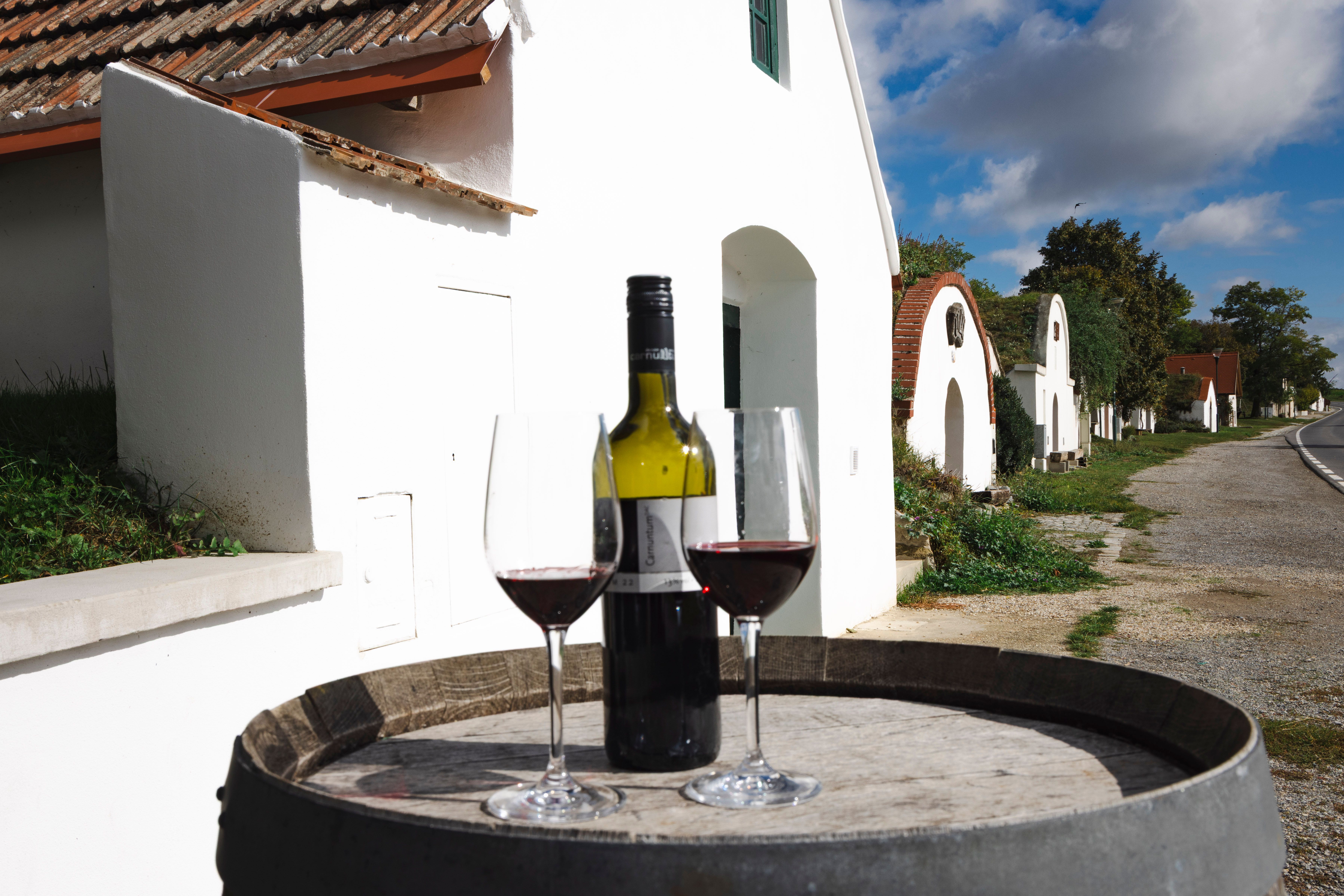 zwei Weingläser und eine Rotweinflasche auf einem Weinfass, im Hintergrund die Kellergasse