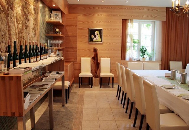 Ein Verkostungsraum mit Holzw&auml;nden, einem langen Tisch mit wei&szlig;en St&uuml;hlen und Weinflaschen auf einem Regal.