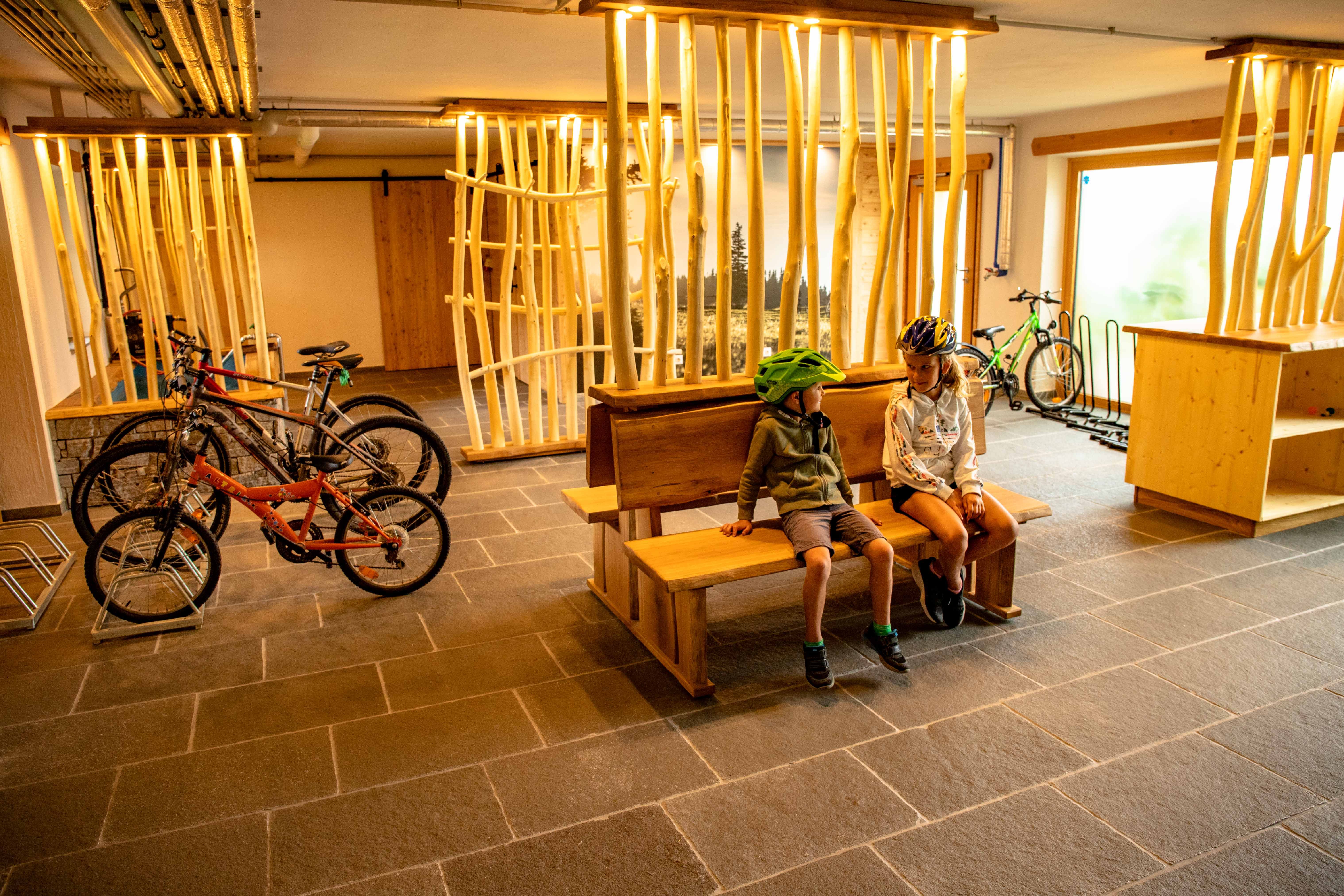 Zwei Kinder mit Fahrradhelmen sitzen auf einer Holzbank in einem Raum mit Fahrrädern und Holzdekor.