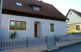 Ein Einfamilienhaus mit grauer Fassade, braunem Dach und Holztüren. Ein kleiner Zaun umgibt das Grundstück.