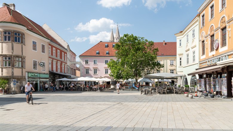 Ein belebter Hauptplatz mit Cafés und Geschäften, umgeben von historischen Gebäuden. Menschen sitzen draußen, ein Radfahrer fährt vorbei. Blauer Himmel mit Wolken.