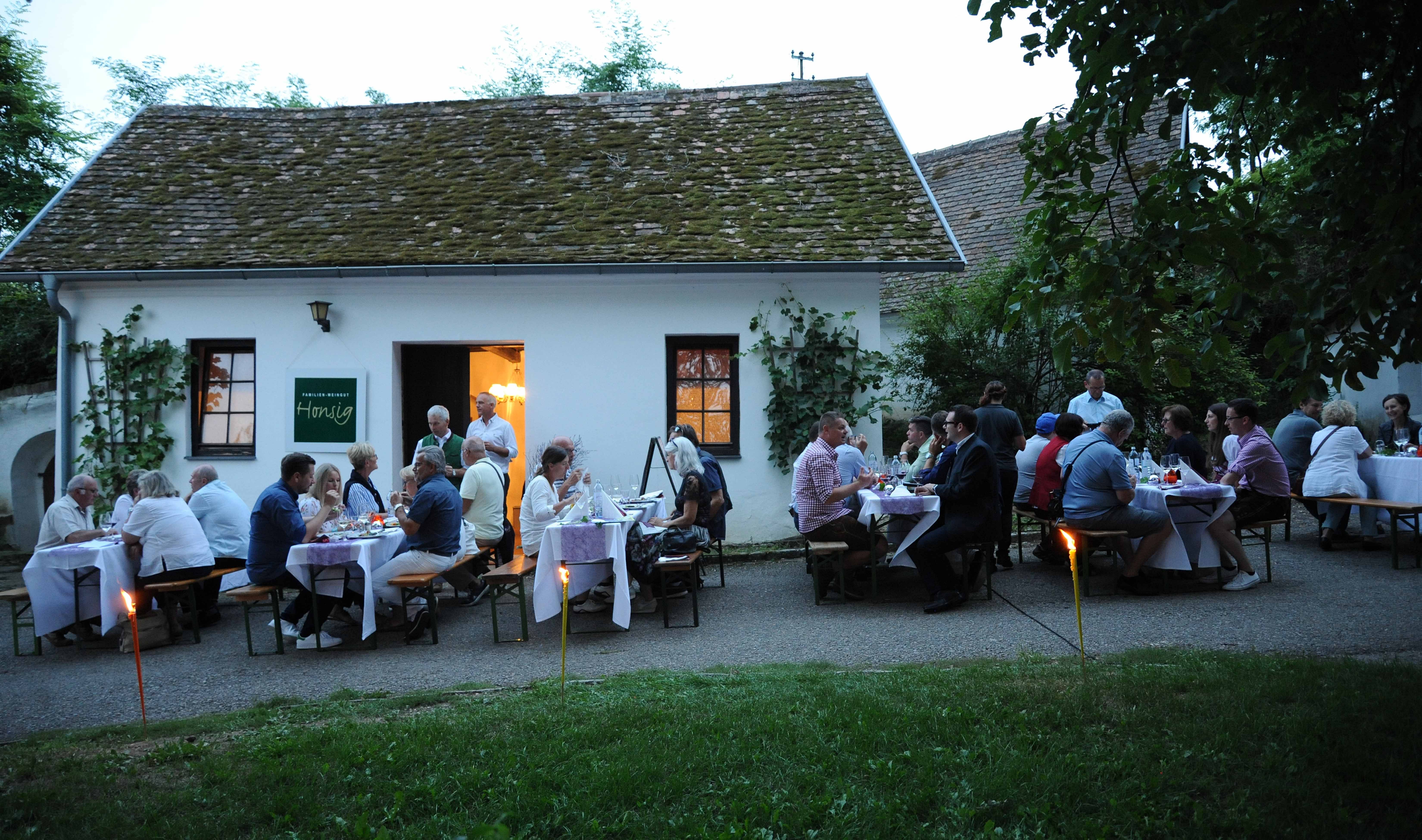 Menschen sitzen abends vor einem Weinkeller und genießen ein Essen im Freien.
