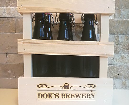 Holzkiste mit drei dunklen Bierflaschen von Dok's Brewery vor einer Steinwand.