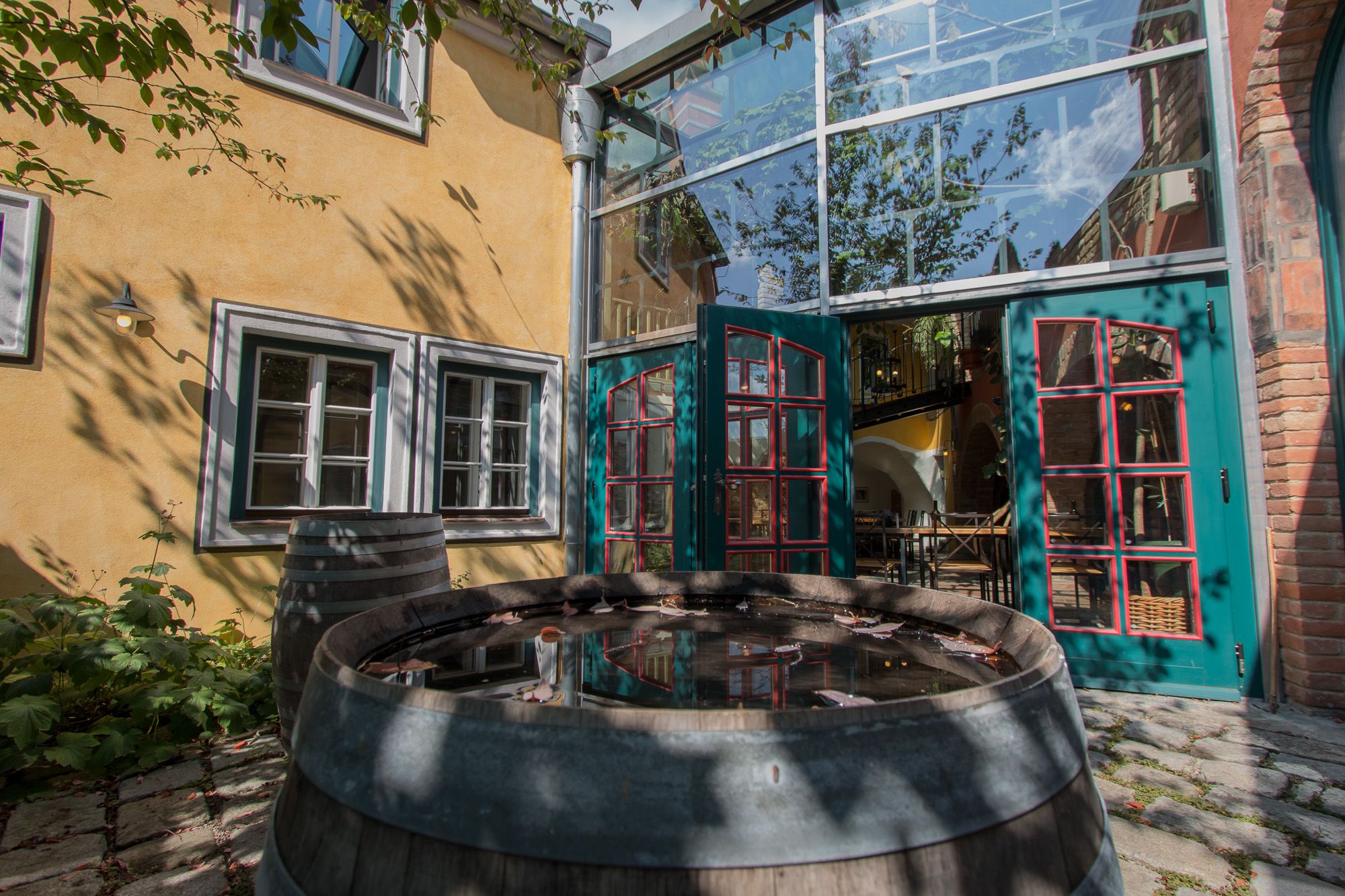 Ein Innenhof mit gelber Fassade, grünen Türen und einem Holzfass mit Wasser. Schatten von Bäumen fallen auf die Wand.