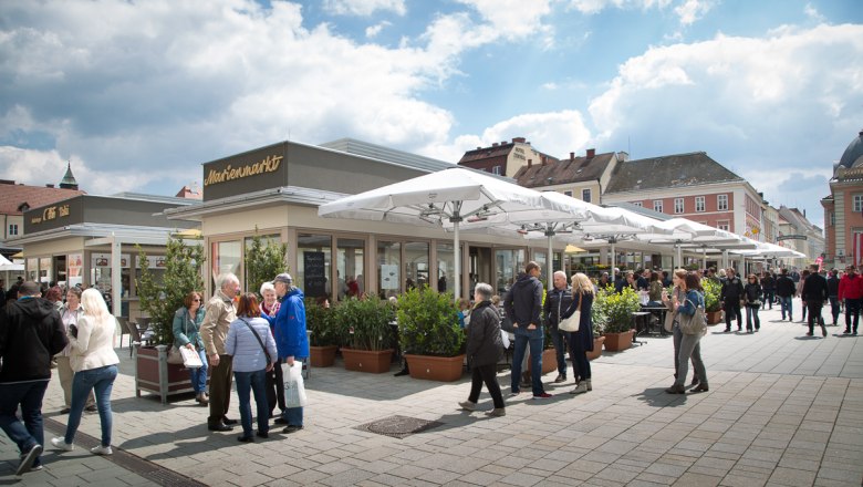 Menschen auf einem belebten Marktplatz mit Cafés und Geschäften unter Sonnenschirmen.