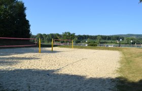 Ein leerer Beachvolleyballplatz mit gelben Pfosten und rosa Netz, umgeben von B&auml;umen und einem Fluss im Hintergrund.