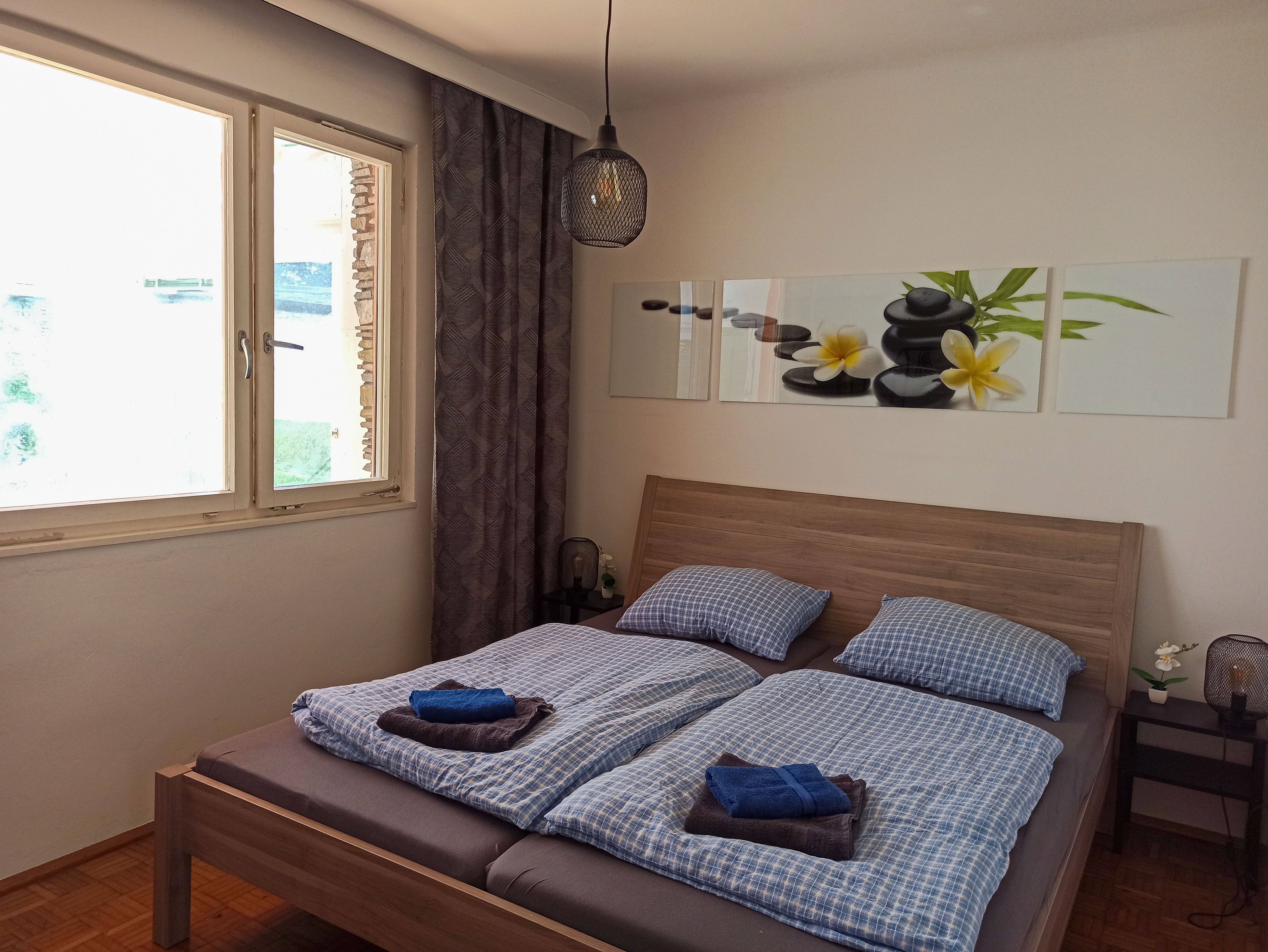 Schlafzimmer mit Doppelbett, blauer Bettwäsche, Bild mit Steinen und Blumen an der Wand, Fenster und Nachttischlampe.