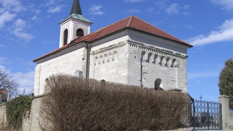 Romanische Kirche mit Glockenturm und roten Ziegeldach, umgeben von einer Mauer und Str&auml;uchern.