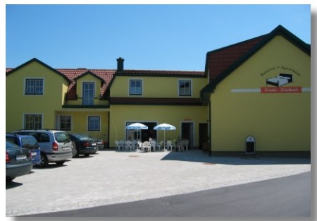 Gelbes Geb&auml;ude mit der Aufschrift 'Pension Ziekel', Autos und Tische mit Sonnenschirmen davor.