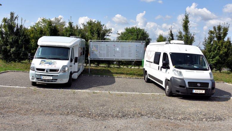 Zwei Wohnmobile auf einem Stellplatz mit Infotafel im Hintergrund.