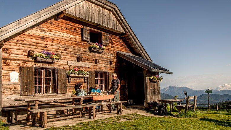 Holzhütte in den Bergen mit Menschen am Tisch, blauer Himmel.