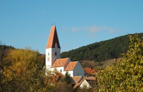 Pfarrkirche Nu&szlig;dorf mit rotem Dach und Uhrturm, umgeben von B&auml;umen und H&uuml;geln.