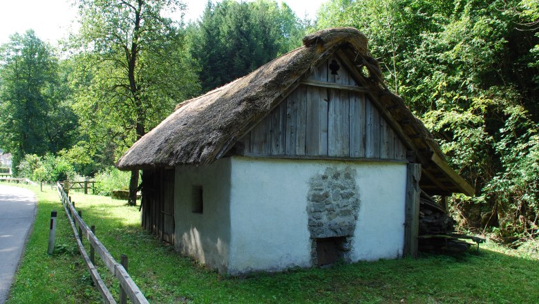 Ein traditionelles D&ouml;rrhaus mit Strohdach in einer l&auml;ndlichen Umgebung, umgeben von B&auml;umen und Wiese.