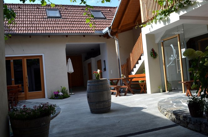 Ein gem&uuml;tlicher Innenhof mit Holzm&ouml;beln, Pflanzen und einem Weinfass. Ein Geb&auml;ude mit roten Dachziegeln und Holztreppe im Hintergrund.