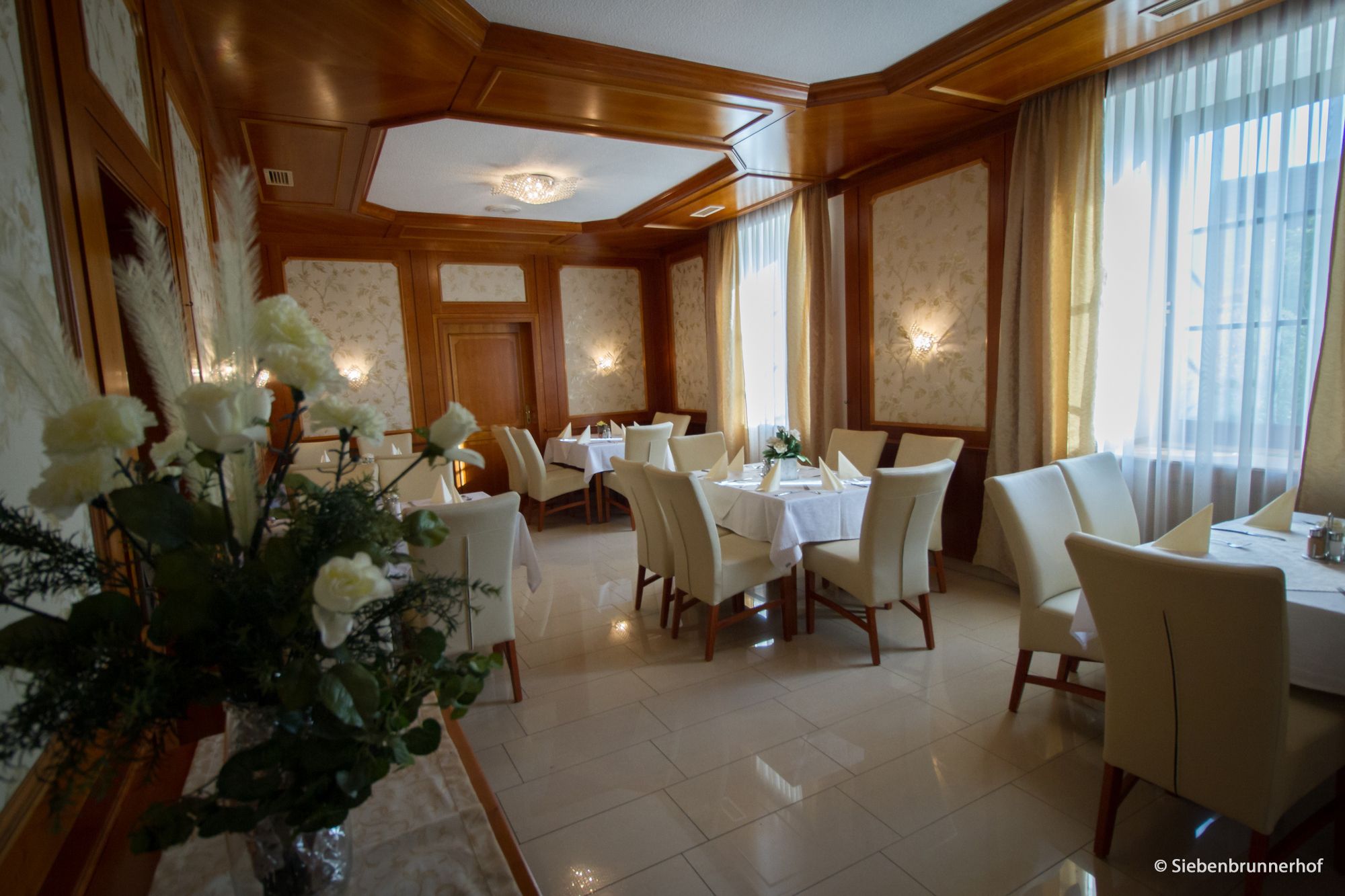 Eleganter Speisesaal mit gedeckten Tischen, weißen Stühlen und Blumenarrangement.