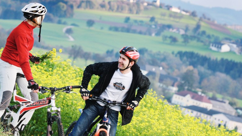 Zwei Radfahrer mit Helmen stehen auf einem bl&uuml;henden Feld mit gr&uuml;ner Landschaft im Hintergrund.