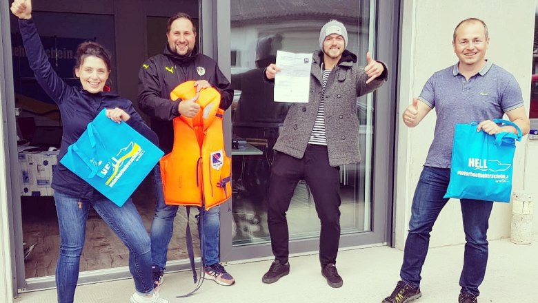 Vier Personen stehen vor einem Geb&auml;ude, halten Taschen und eine Schwimmweste, l&auml;cheln und zeigen Daumen hoch.