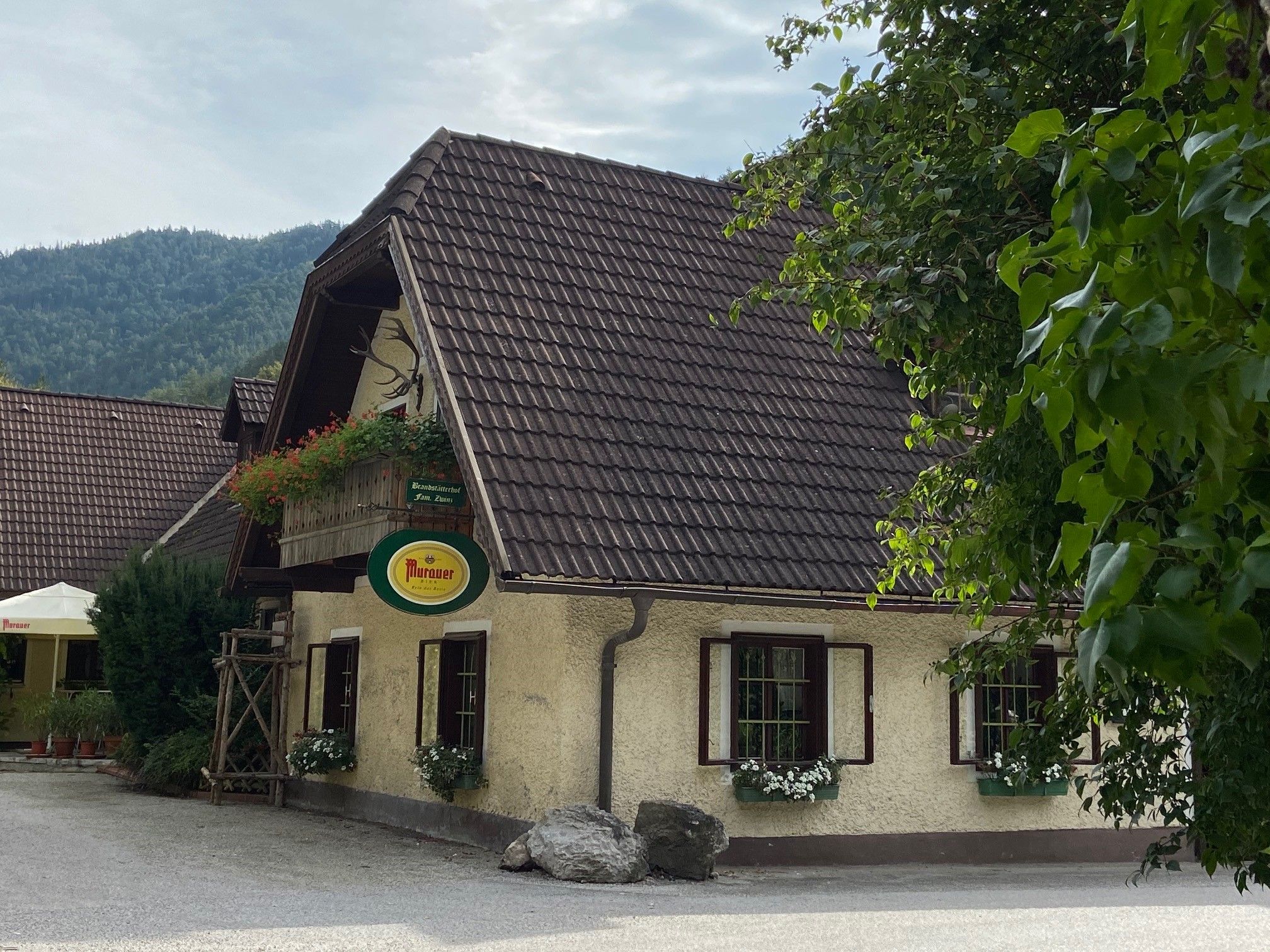 Traditionelles Gasthaus mit Blumen und Murauer-Schild, umgeben von Bäumen.