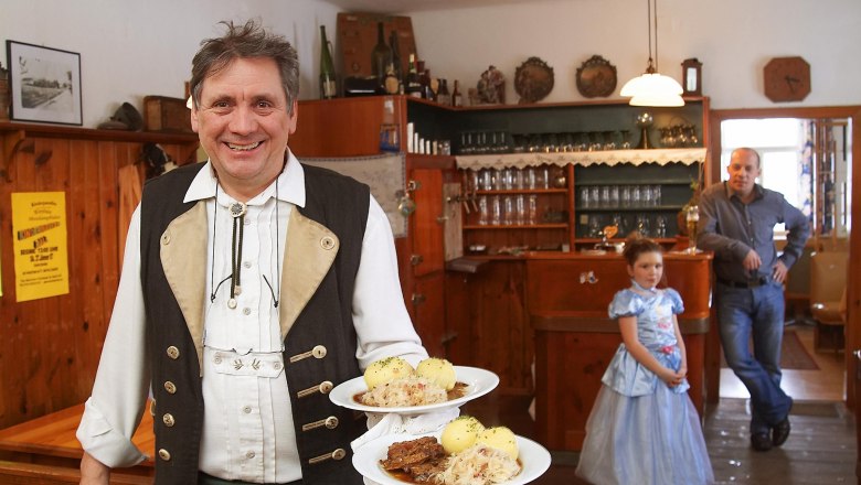 Ein Mann in traditioneller Kleidung serviert Schweinsbraten mit Kn&ouml;deln in einem rustikalen Gasthaus. Im Hintergrund ein Mann und ein M&auml;dchen im Kleid.