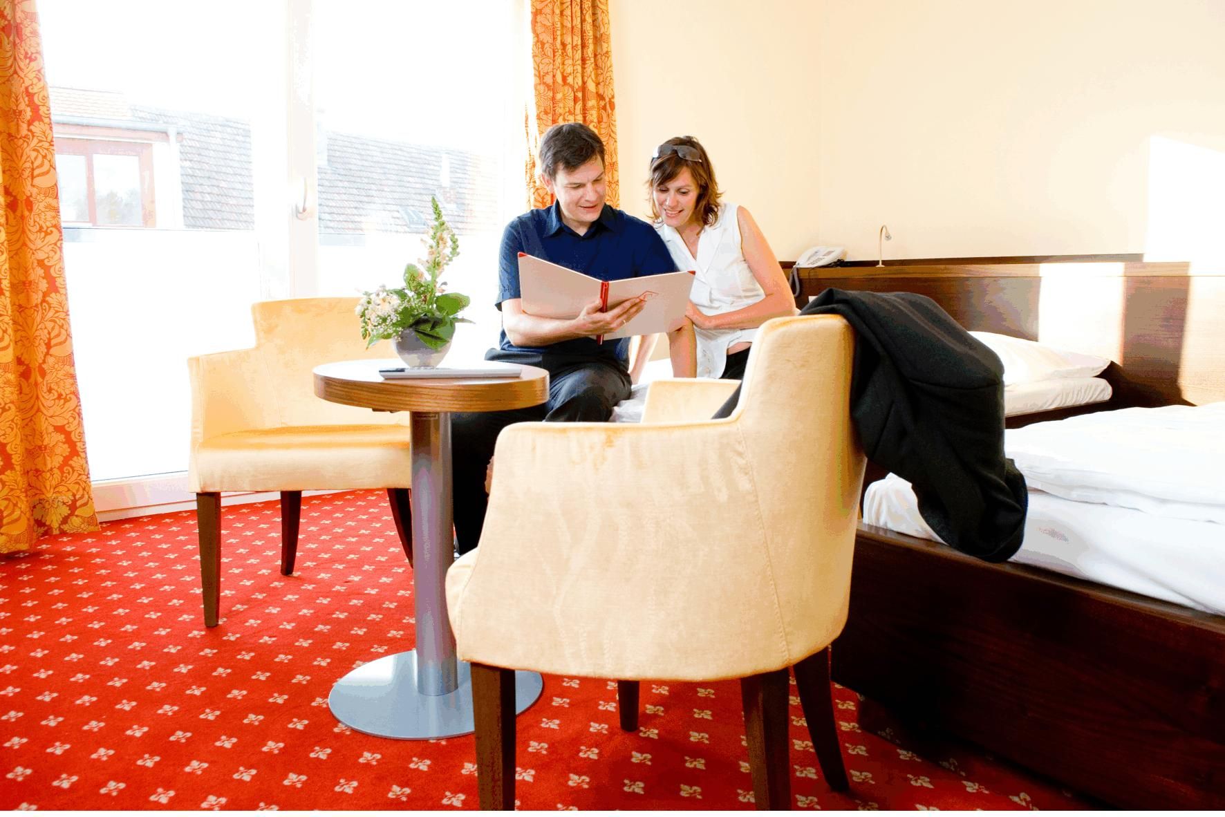 Ein Paar sitzt in einem Hotelzimmer auf einem Bett und schaut in ein Buch. Der Raum hat rote Teppiche und gelbe Vorhänge.