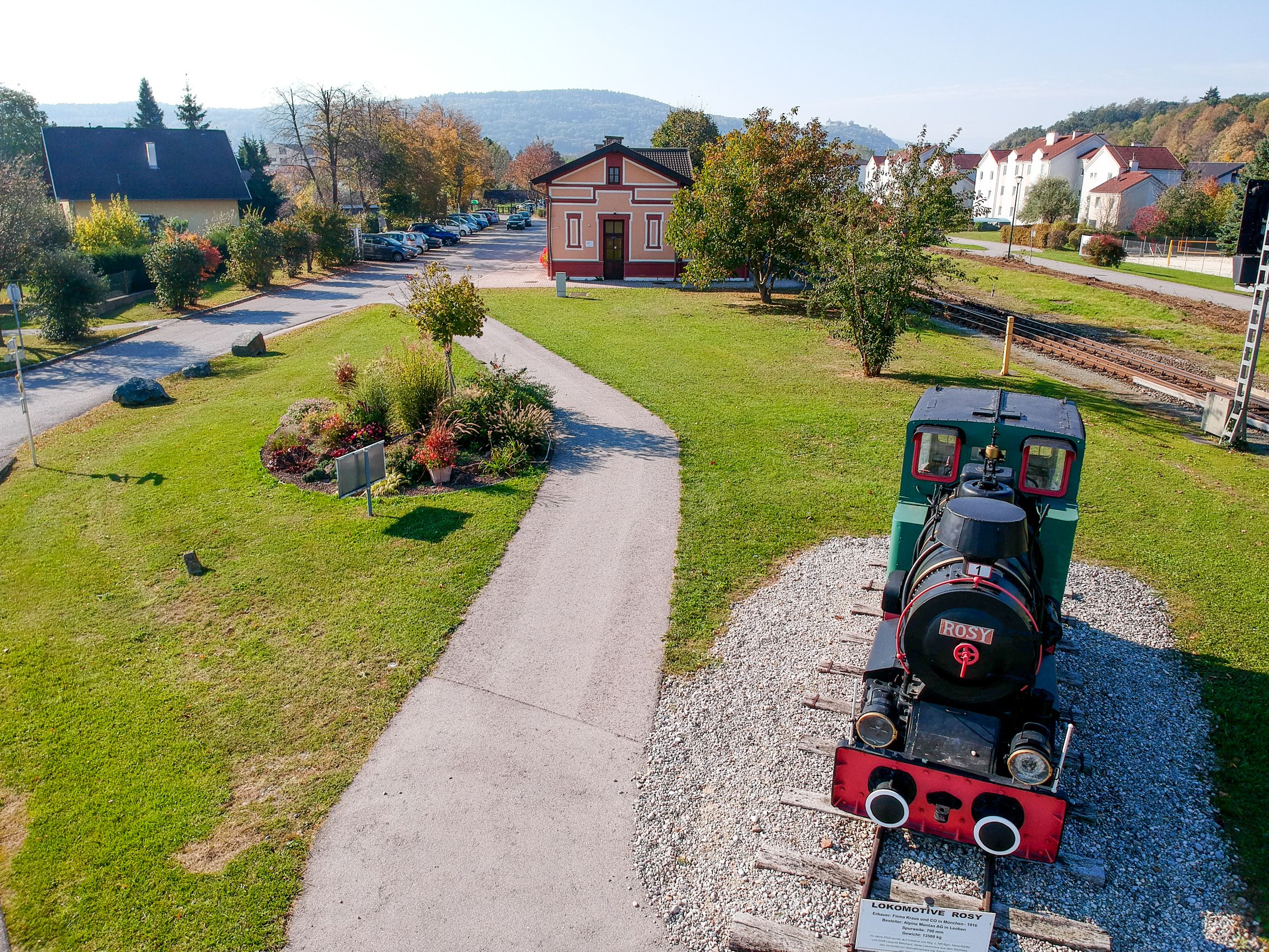 Eine kleine historische Lokomotive namens 'Rosy' steht auf einem kurzen Gleisabschnitt in einem grünen Park neben einem kleinen Bahnhofsgebäude.
