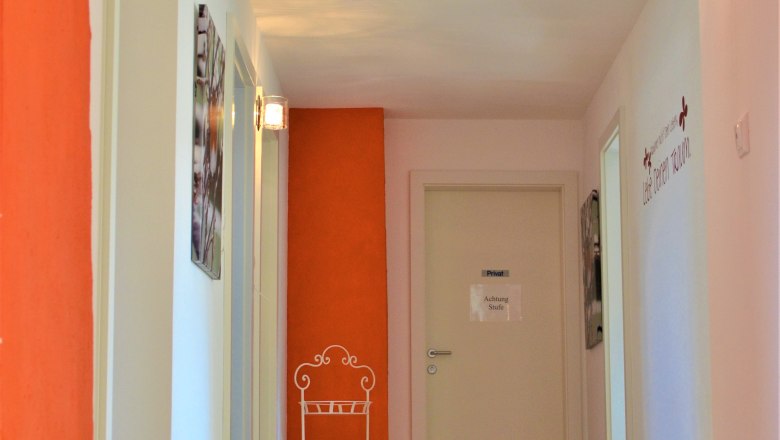 Ein Flur mit Holzboden, weißen Wänden und einer orangefarbenen Wand. An der Wand hängt ein Schild mit der Aufschrift 'Achtung Stufe'.