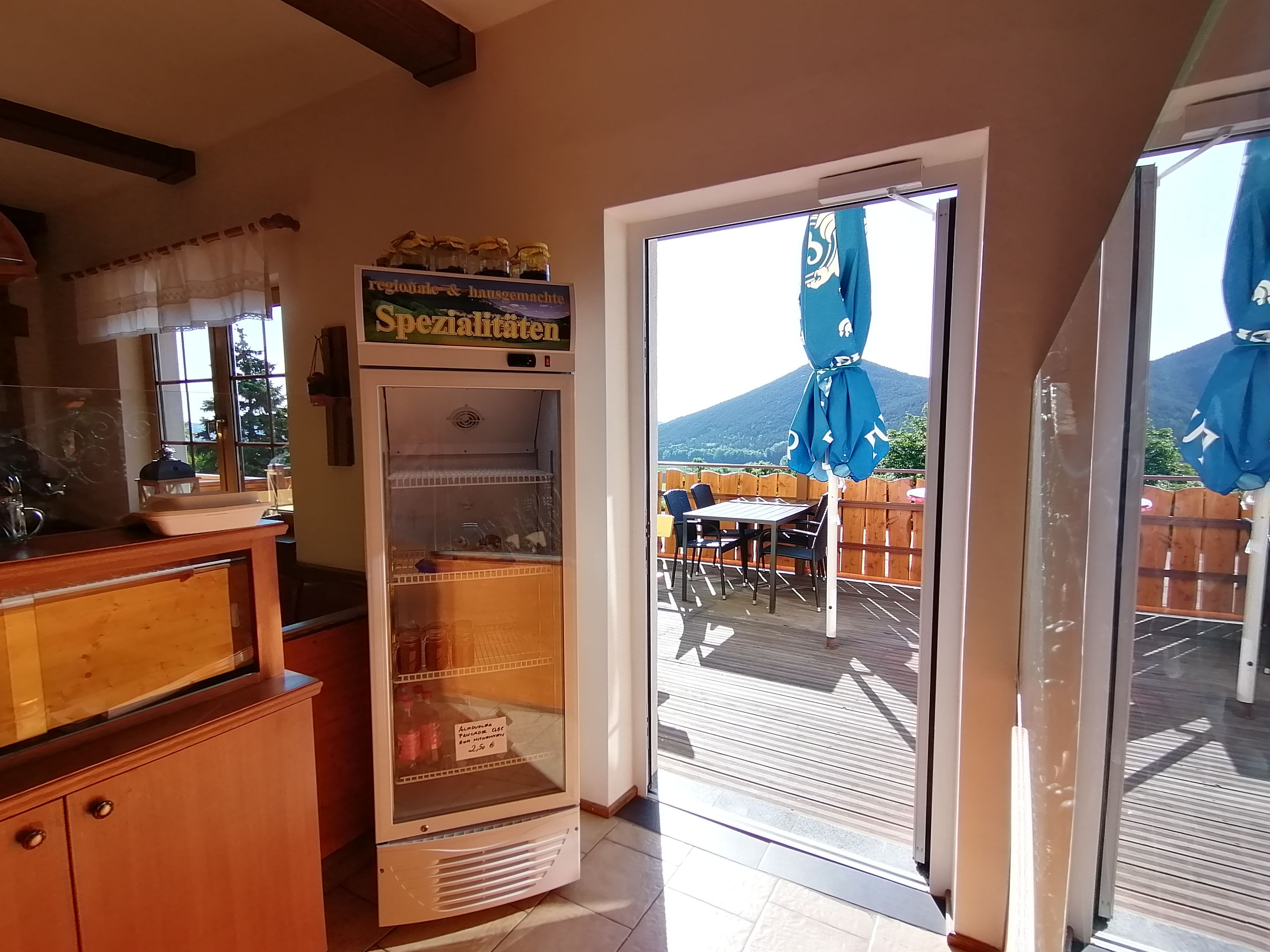 Blick von innen auf eine Terrasse mit Sonnenschirmen und Bergen im Hintergrund.