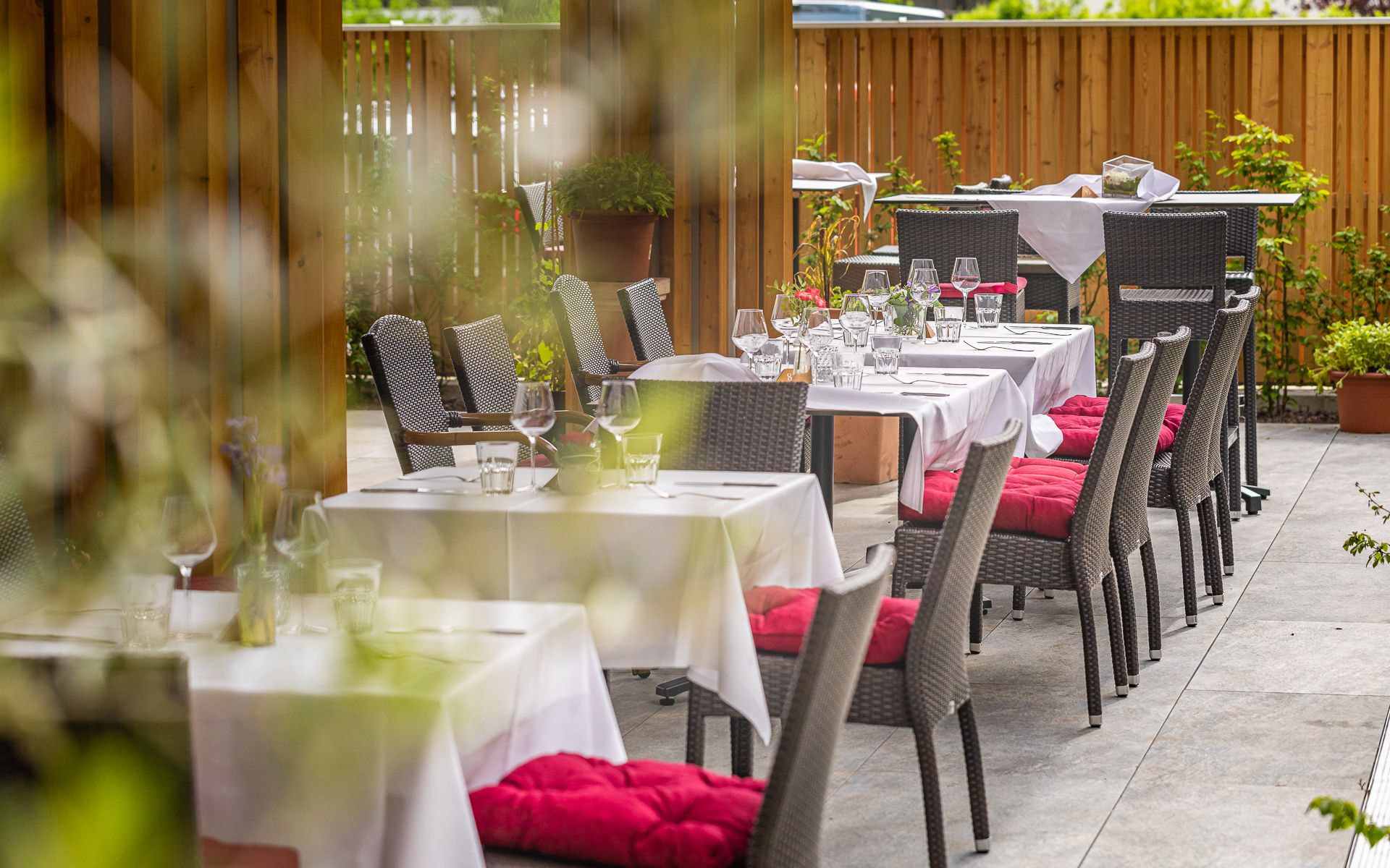 Gedeckte Tische auf einer Restaurantterrasse mit roten Sitzkissen und Holzzaun im Hintergrund.