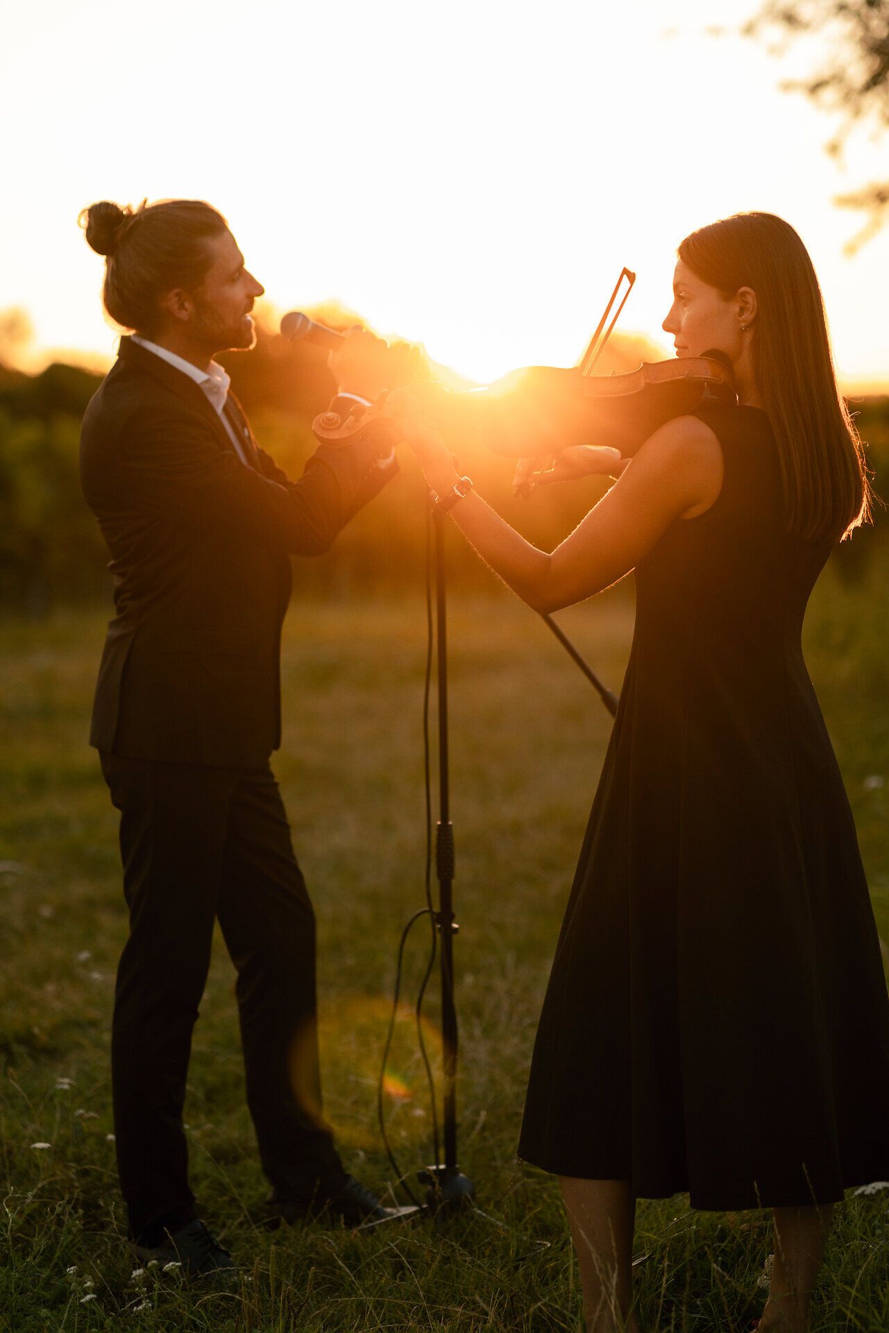 Eine Violinistin und ein Violinist spielen im Freien bei Sonnenuntergang. Die tief stehende Sonne scheint zwischen den beiden Musizierenden hindurch.